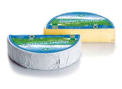 KI generiert: Das Bild zeigt ein Stück weißen Käse, das auf einer schwarzen Schieferplatte in vier Portionen geschnitten ist, daneben ist eine verpackte, identische Käserolle abgebildet.