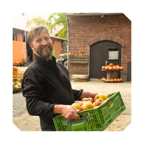 KI generiert: Ein Mann hält draußen eine Kiste mit Äpfeln. Im Hintergrund sind Kürbisse zu sehen.