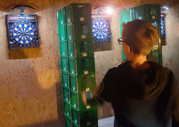 KI generiert: Ein Junge spielt Darts in einem Raum mit grünen Kisten.
