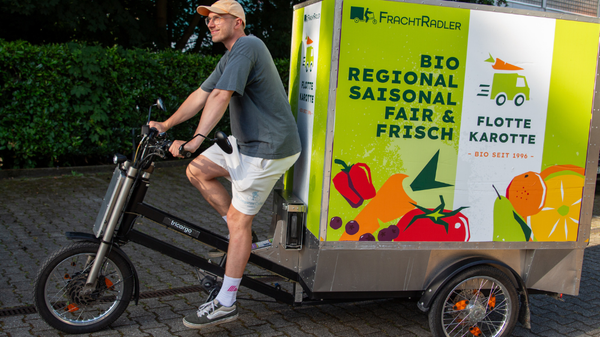 KI generiert: Ein Mann fährt ein Lastenrad mit Werbung für "Flotte Karotte", Bio-Obst und Gemüse. Text: "BIO REGIONAL SAISONAL FAIR FRISCH".