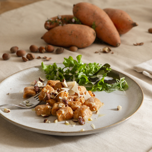 KI generiert: Ein Teller mit Gnocchi, Salat und Nüssen, im Hintergrund Süßkartoffeln und Haselnüsse.