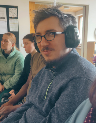 KI generiert: Eine Person mit Brille und Gehörschutz in einem Raum mit mehreren Menschen.