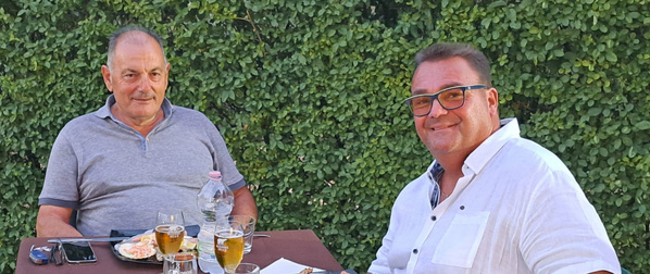 KI generiert: Zwei Männer sitzen an einem Tisch mit Getränken und Essen im Freien vor einer grünen Hecke.