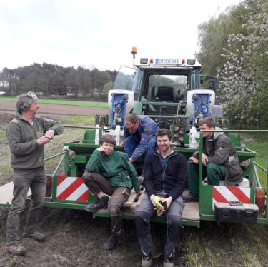 KI generiert: Gruppe von fünf Personen sitzt und steht auf einem landwirtschaftlichen Anhänger vor einem Traktor auf einem Feld.