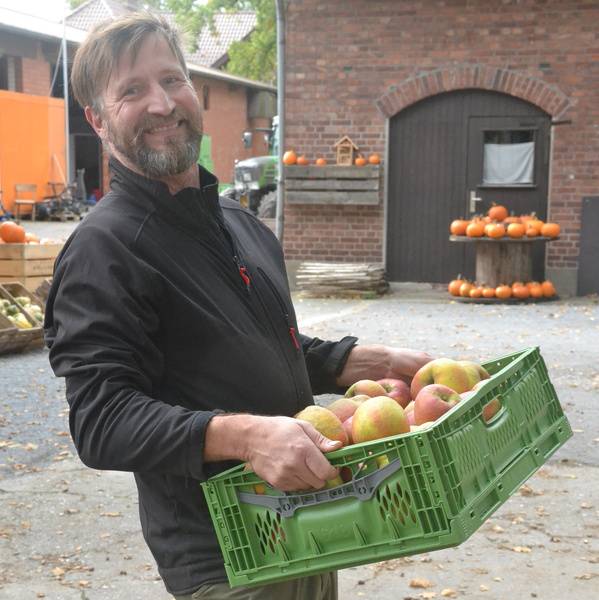 KI generiert: Ein Mann lächelt und hält eine Kiste voller Äpfel in einem Hof mit Kürbissen im Hintergrund.
