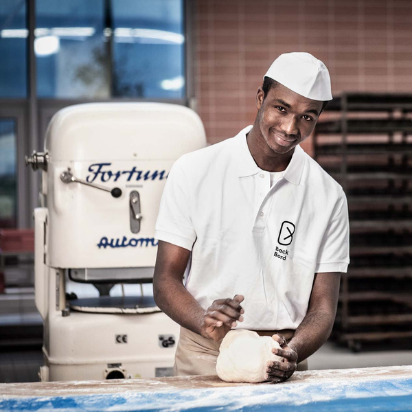 KI generiert: Ein Bäcker formt Teig mit der Hand, im Hintergrund eine Maschine. Auf dem Hemd steht "Back Bord".