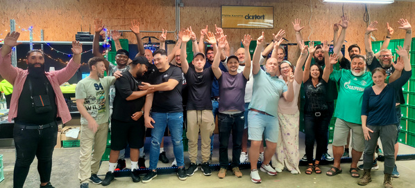 KI generiert: Eine Gruppe von Menschen posiert fröhlich in einer Lagerhalle mit grünen Kisten im Hintergrund. 