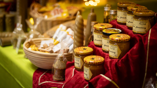 KI generiert: Honiggläser auf einem Tisch mit roten Tüchern, im Hintergrund Körbe mit Wachs und Kerzen. Text: "Bienenhonig".