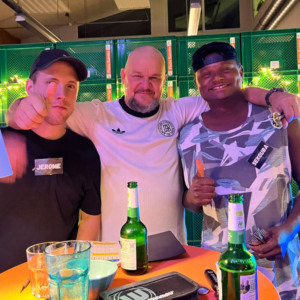 KI generiert: Drei Personen posieren fröhlich an einem Tisch mit Getränken. Text: "JEROME", "SEKOUBA" auf Namensschildern.