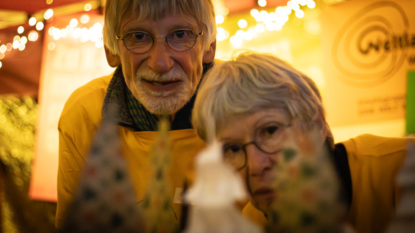 KI generiert: Zwei ältere Personen in gelben Hemden bei einem Weihnachtsmarktstand. Text im Hintergrund unklar.