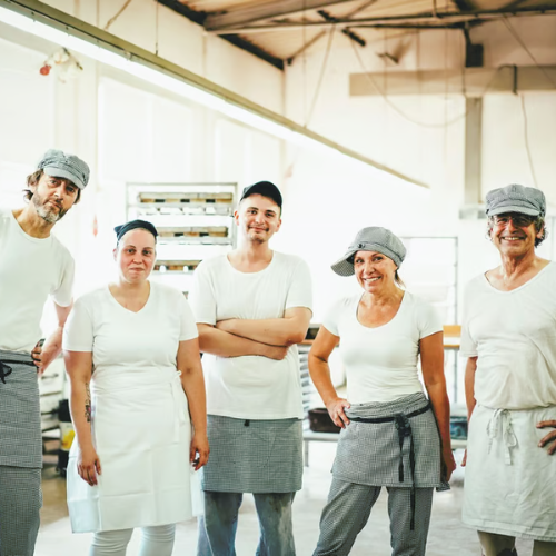 KI generiert: Eine Gruppe von fünf Bäckern in weißer Arbeitskleidung posiert und lächelt in einer Bäckerei.