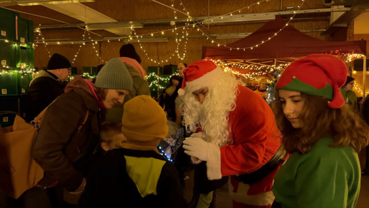 KI generiert: Auf dem Bild ist eine weihnachtliche Szene mit einem Weihnachtsmann zu sehen, der mit Kindern spricht. Im Hintergrund sind Menschen, dekorative Lichter und ein Pavillon zu erkennen.