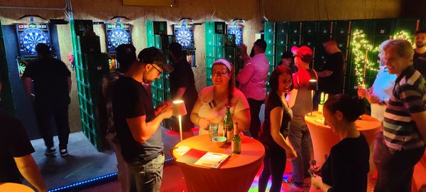 KI generiert: Eine Gruppe spielt in einer Bar Darts; Leute unterhalten sich an Stehtischen mit Getränken.