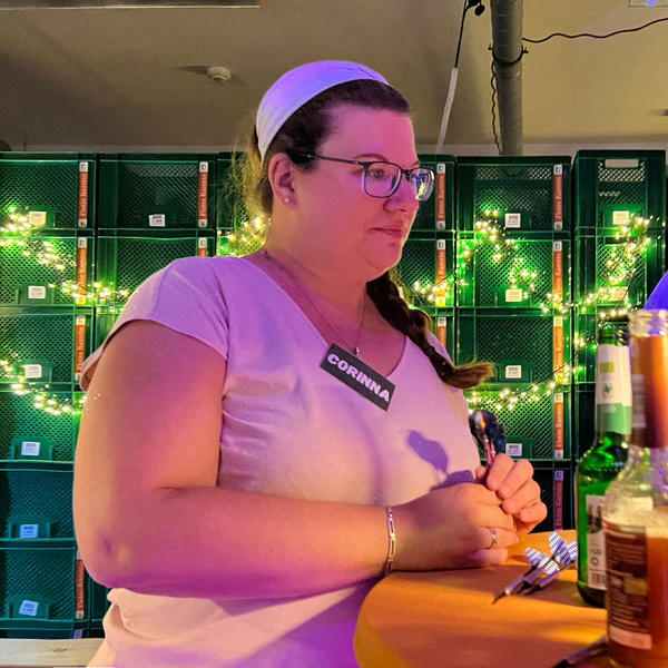 KI generiert: Eine Person steht an einem Tisch mit Getränken vor grünen Kisten und Lichterketten. Namensschild: "CORINNA".